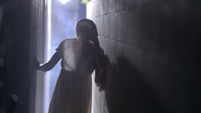 A backlit woman in a nightgown steadies herself between two walls in a narrow alley.