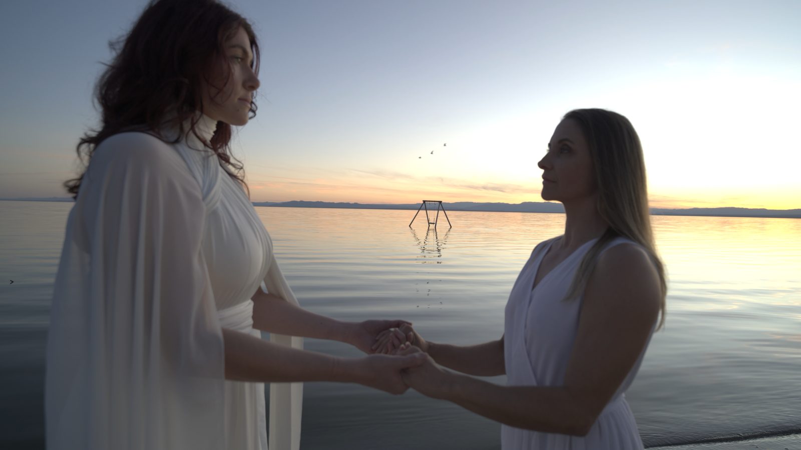 Artemis: two women in white hold hands at the water's edge at sunset, a swing set standing in the lake behind them.