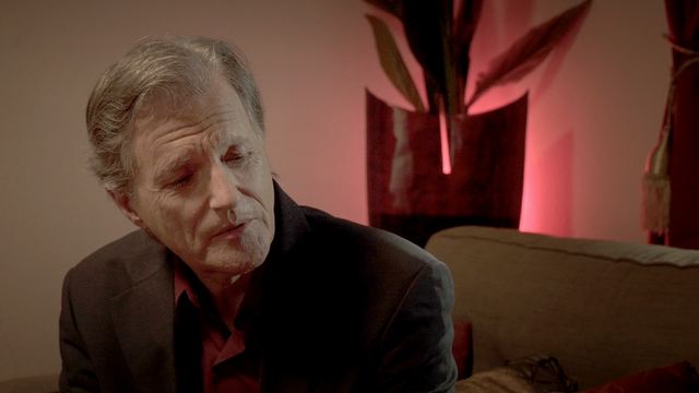 An older man looks down, a red-lit vase behind him.
