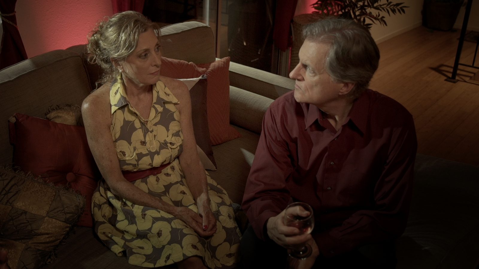 Black King Down: a woman sits on a couch beside a man holding a glass of wine, both tense, lit by red lamps.