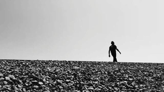 A lone figure walking along a gravel ridge against a white sky.