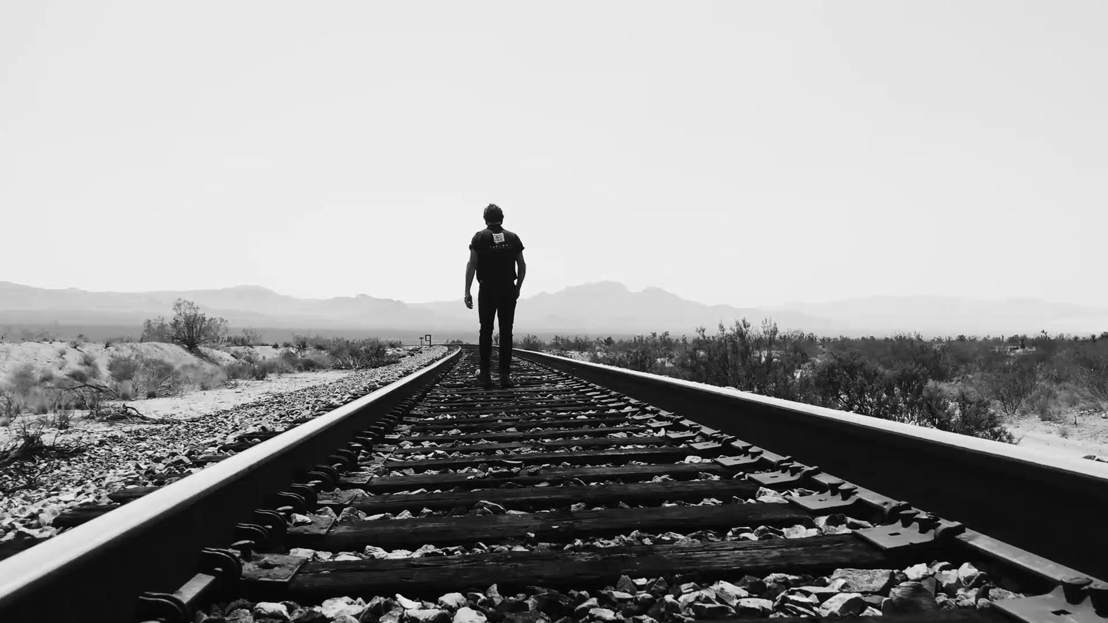 My Hands: a man walks away down desert railroad tracks toward distant mountains, in black and white.