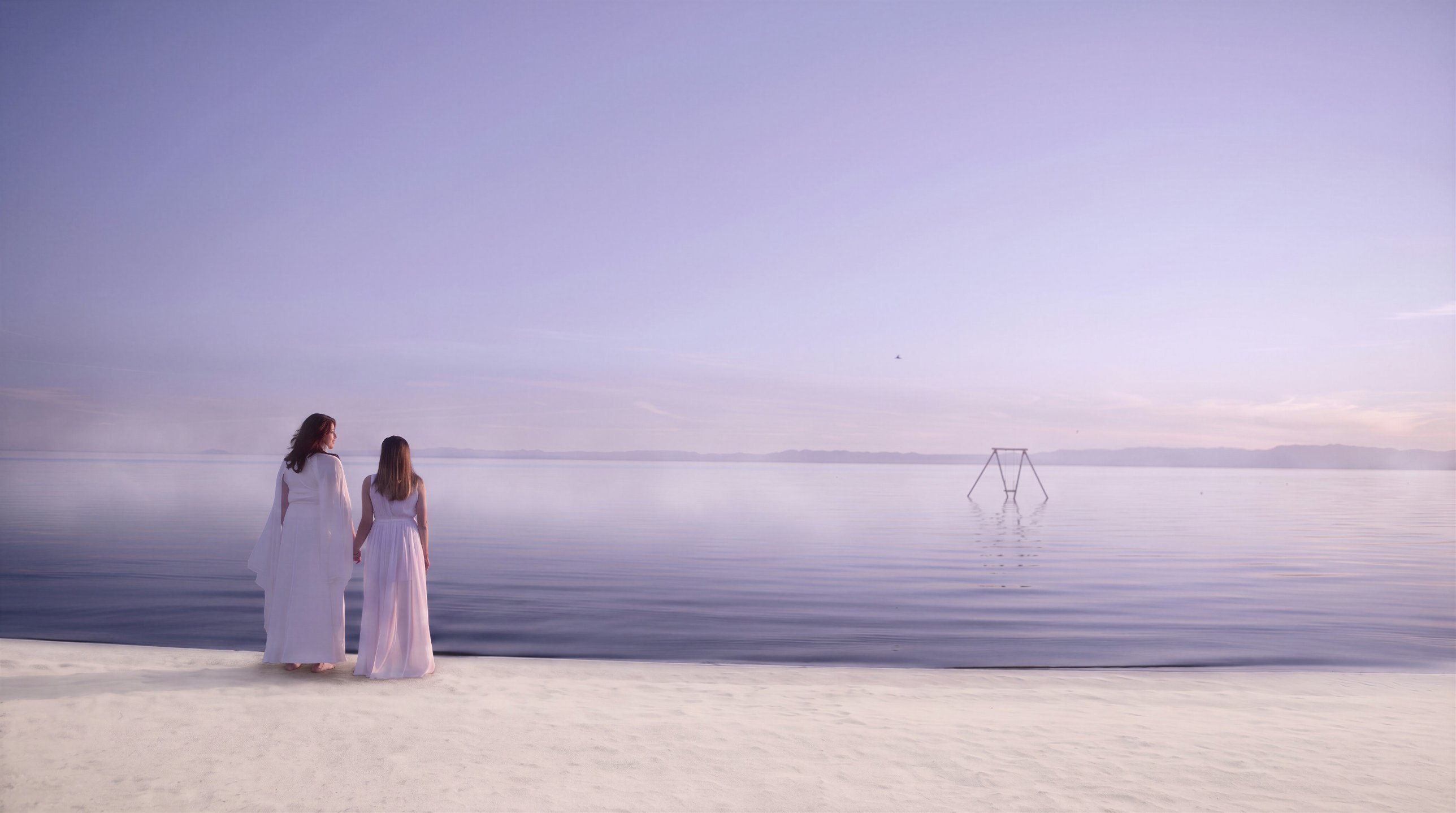 Two women in white stand hand in hand on a pale white shore, looking out over a still lavender lake toward an empty swing set.